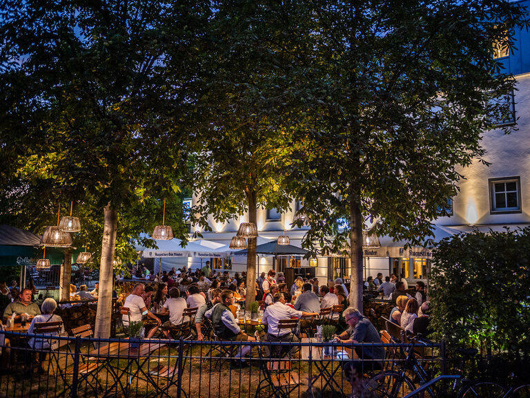 Menschen genießen den Abend in dem Biergarten des Toni´s Gasthaus unter Bäumen.