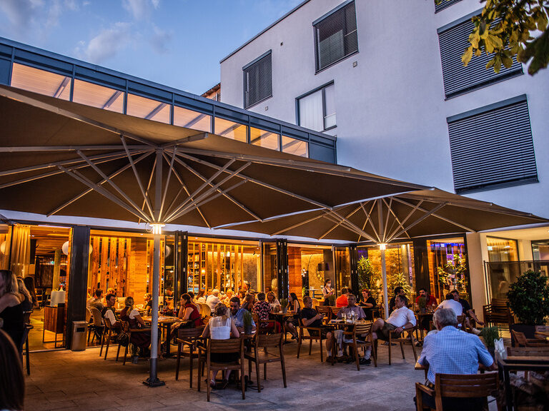 Eine fröhliche Gruppe unter Sonnenschirmen im Biergarten des Toni´s Gasthaus by Genusshotel Wenisch.