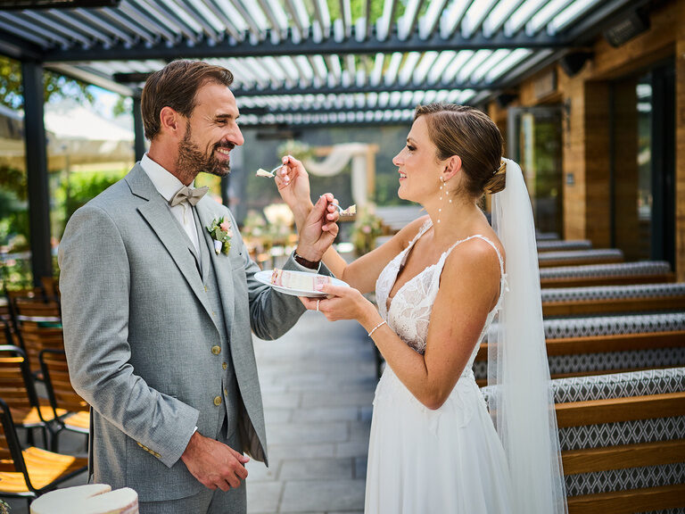 Hochzeitspaar teilt sich Torte und lacht bei Feier im modernen Outdoor-Ambiente