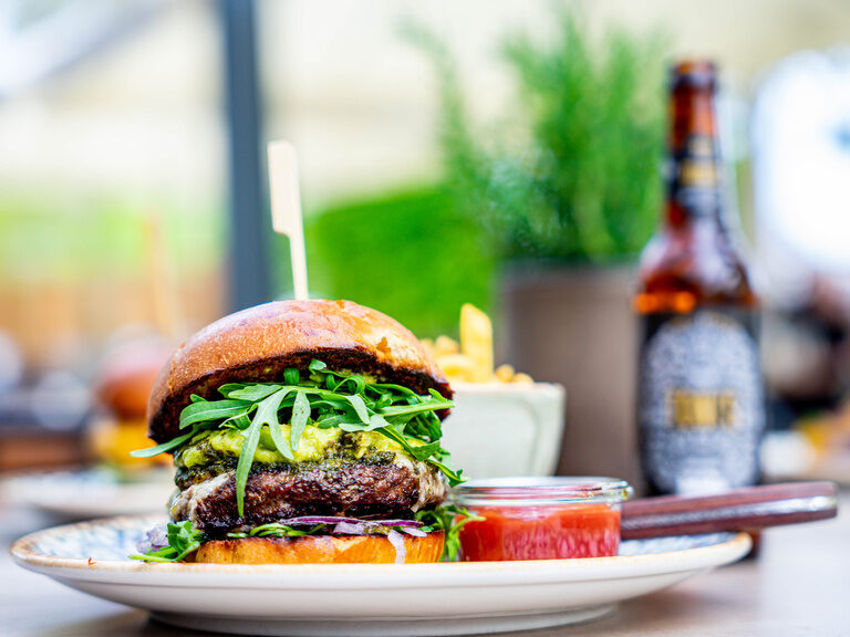 Ein Hamburger auf einem Teller im Genusshotel Wenisch, mit einer Bierflasche im Hintergrund.