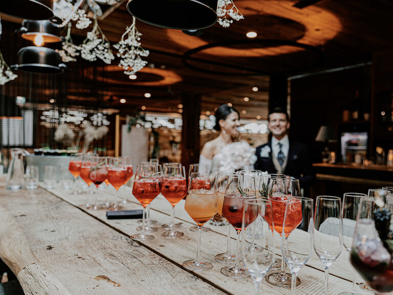 Eine große Auswahl an Cocktails steht auf einem Holztisch umgeben von Menschen in schicken Gewänder