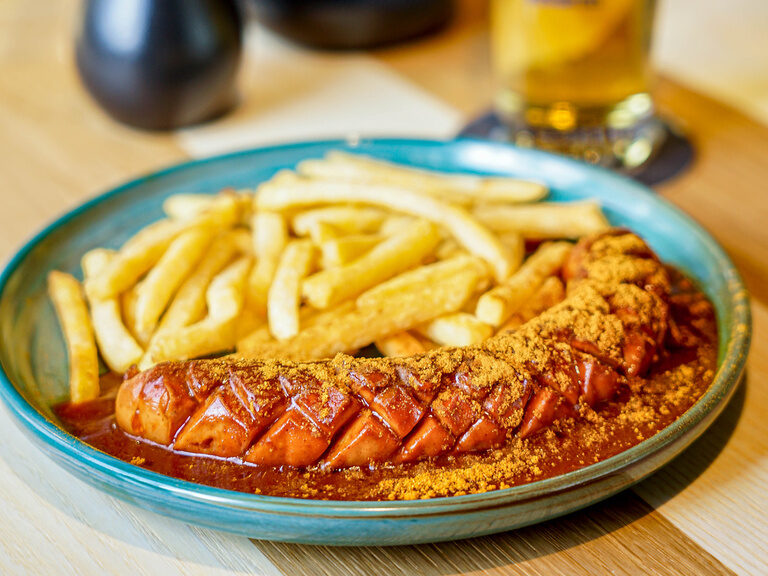 Auf einem Teller ruht eine Currywurst neben Pommes und ein Bier steht daneben.