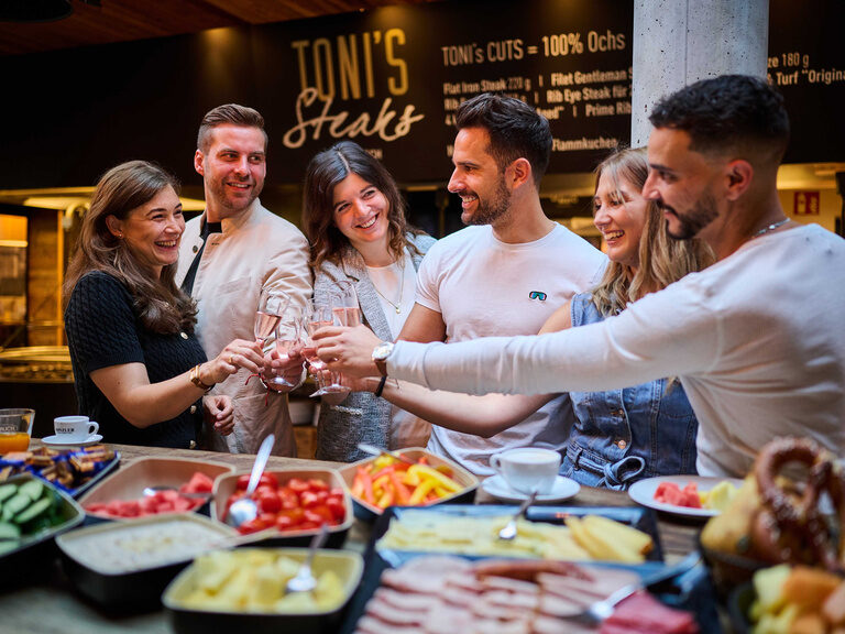 Ein fröhlicher Frühstückstisch im Restaurant mit sechs lächelnden Personen in entspannter Kleidung.