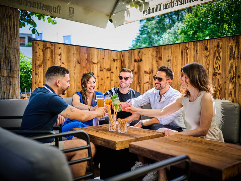 Eine Freundesgruppe stößt mit ihren Gläsern auf einer Terrasse an einem Holztisch an.