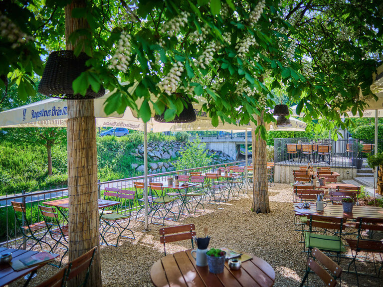 Ein gemütliches Biergarten-Szenario im Genusshotel Wenisch mit Tischen und Stühlen unter einem schattenspendenden Baum.
