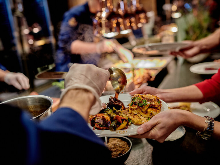 Eine Person verteilt Soße auf dem Teller einer anderen Person, welcher mit Essen gefüllt ist.