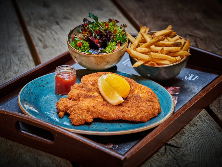 Ein saftiges Schnitzel liegt auf einem Teller neben Pommes und einem bunten Salat.